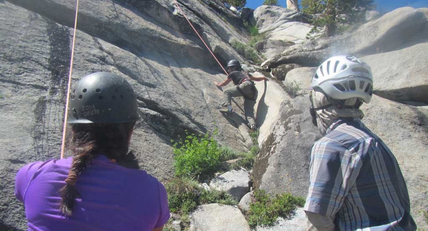 two people wearing helmets look up as another person rock climbs above
